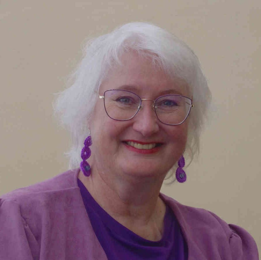 The author Sheila Crosby. She has short white hair and pale skin. She's wearing a purple top, a lilac blouse and dangly lilac earings.